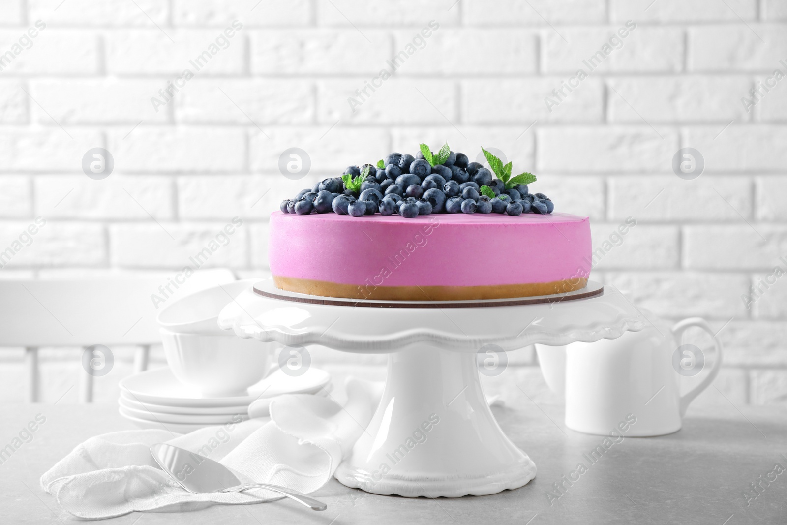 Photo of Stand with tasty blueberry cake on grey table against white brick wall