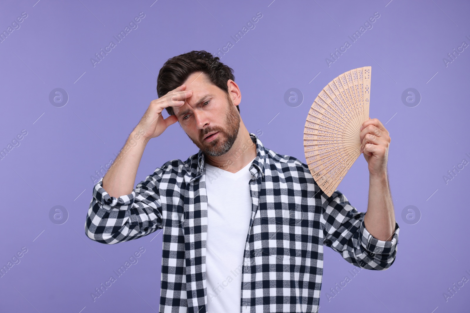 Photo of Unhappy man with hand fan suffering from heat on purple background