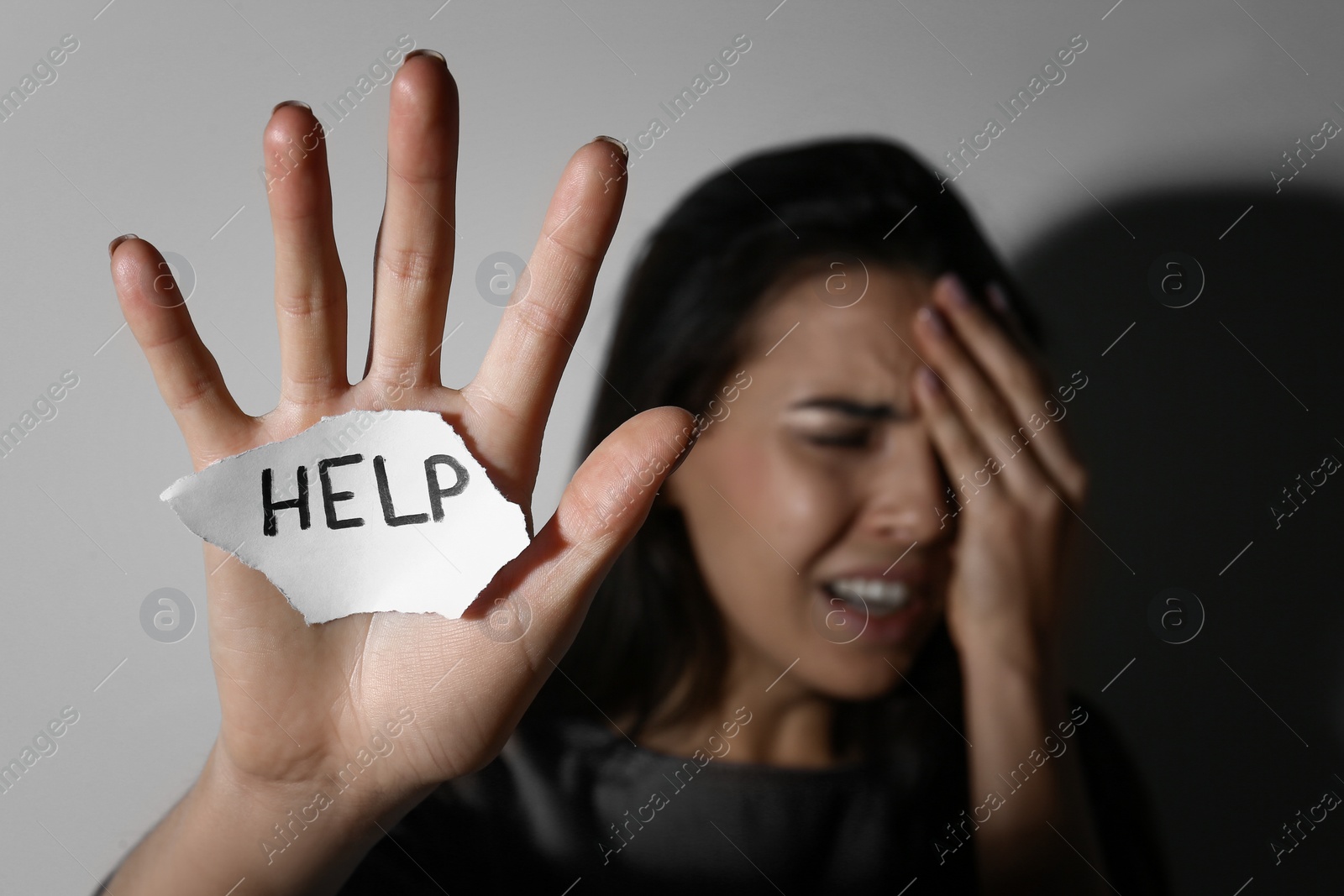 Photo of Crying young woman with sign HELP near white wall, focus on hand. Domestic violence concept