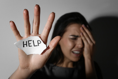 Photo of Crying young woman with sign HELP near white wall, focus on hand. Domestic violence concept