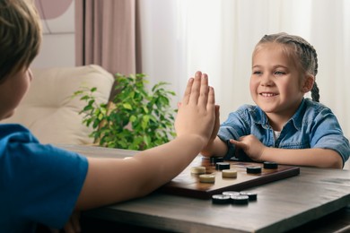 Playing checkers. Cute boy giving high five to little girl at table in room, closeup