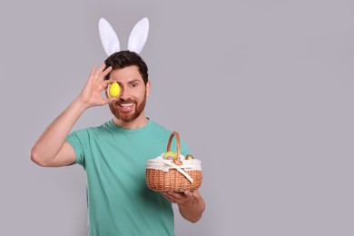 Happy man in cute bunny ears headband covering eye with Easter egg and holding wicker basket on light grey background. Space for text