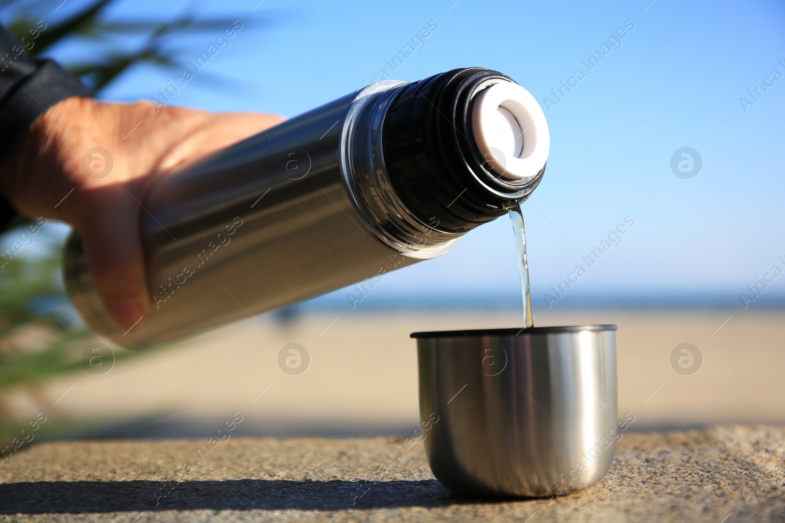 Photo of Woman pouring hot drink from metallic thermos into cap on stone surface outdoors, closeup