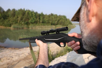Photo of Man aiming with hunting rifle near lake outdoors, closeup