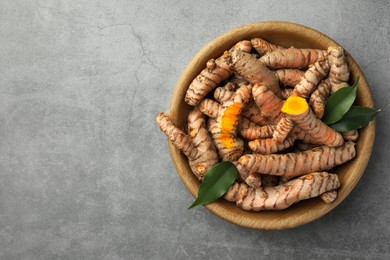 Photo of Bowl with raw turmeric roots and green leaves on grey table, top view. Space for text