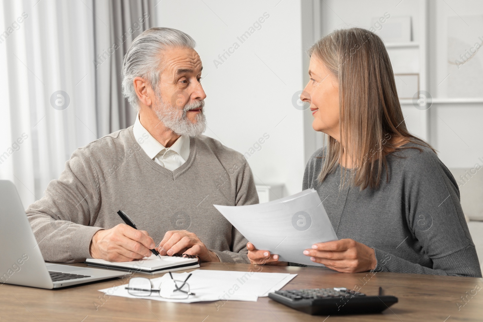 Photo of Elderly couple with papers and laptop discussing pension plan at wooden table in room