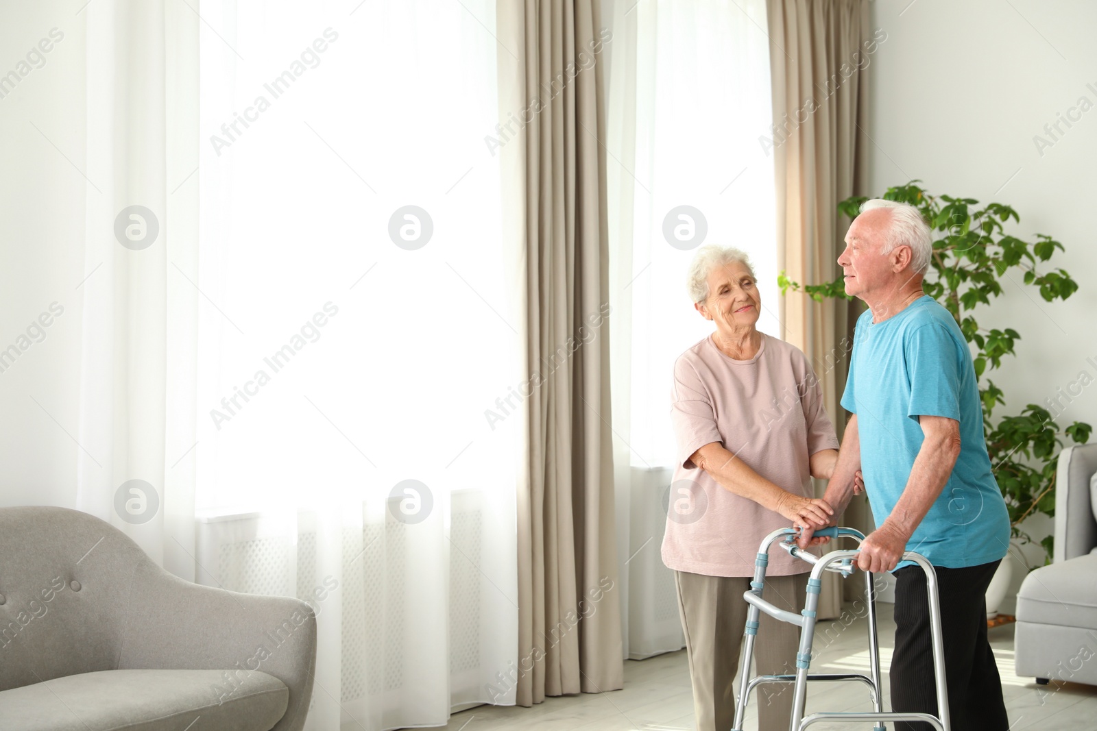 Photo of Elderly woman and her husband with walking frame indoors. Space for text