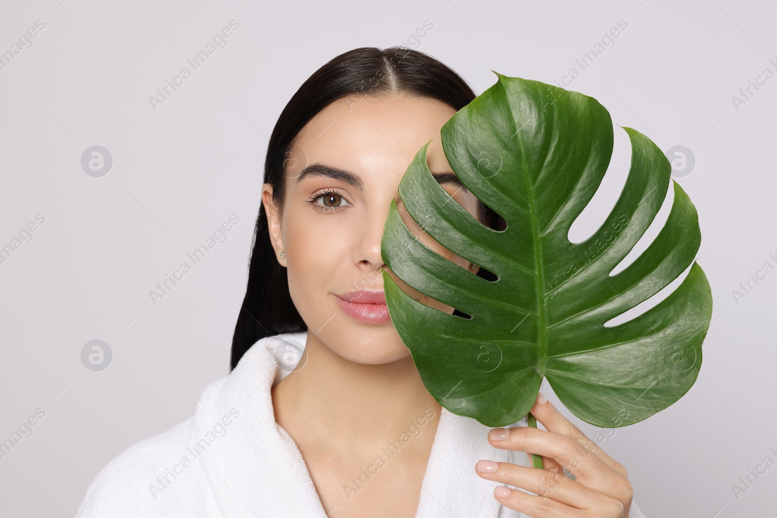 Photo of Woman in bathrobe holding leaf of monstera on light grey background. Spa treatment