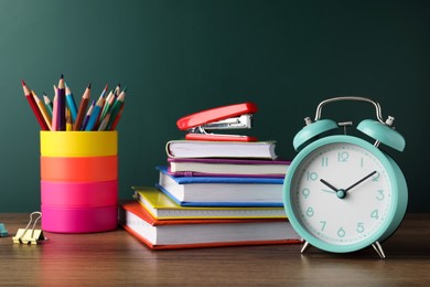 Photo of Alarm clock and different stationery on wooden table near green chalkboard. School time