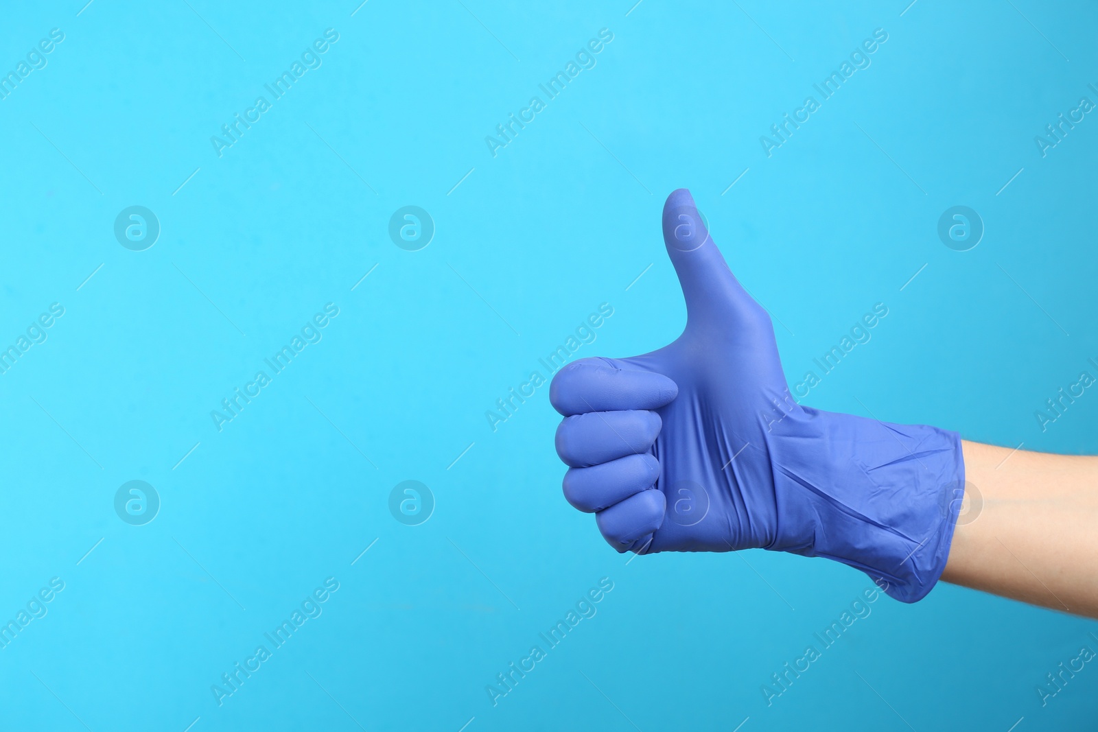 Photo of Person in medical gloves showing thumb up on light blue background, closeup of hand. Space for text