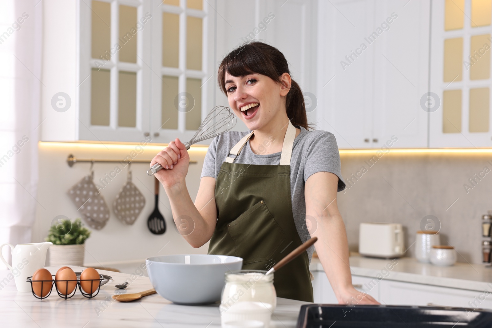 Photo of Happy young housewife with whisk having fun while cooking at white marble table in kitchen
