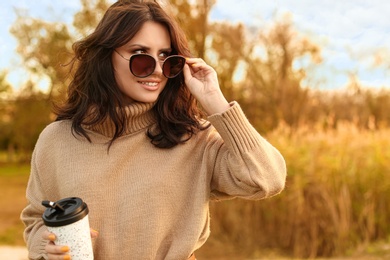 Beautiful young woman with cup of coffee wearing stylish autumn sweater outdoors