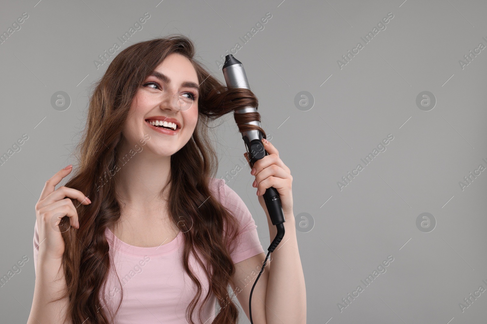 Photo of Beautiful young woman using curling hair iron on grey background, space for text