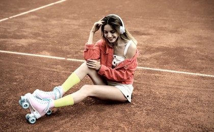 Image of Happy stylish young woman with vintage roller skates and headphones sitting on tennis court