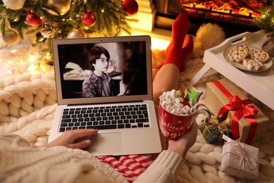 Photo of MYKOLAIV, UKRAINE - DECEMBER 23, 2020: Woman with sweet drink watching Harry Potter and Philosopher's Stone movie on laptop near fireplace at home, closeup. Cozy winter holidays atmosphere