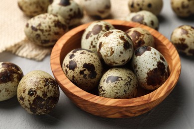Photo of Wooden bowl and many speckled quail eggs on grey table, closeup