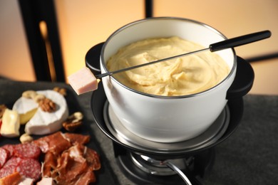 Photo of Fork with piece of ham, fondue pot with melted cheese and snacks on table, closeup