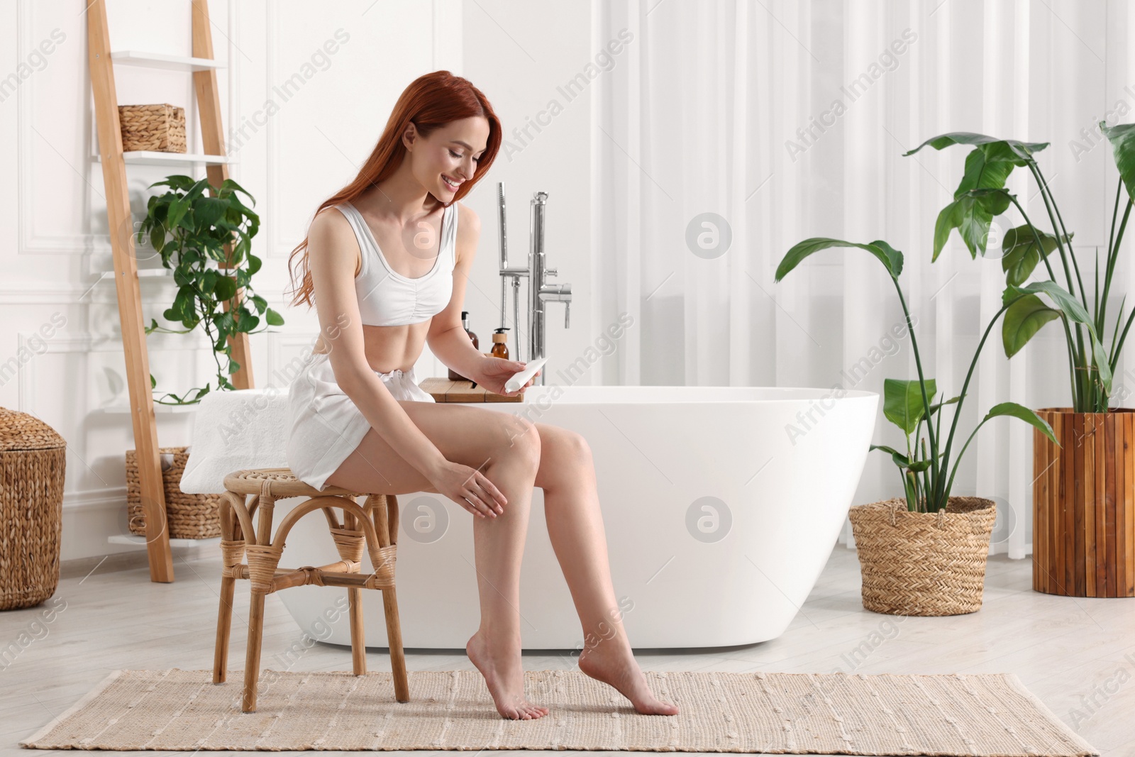Photo of Beautiful young woman applying body cream onto legs in bathroom, space for text
