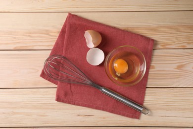 Photo of Metal whisk, raw eggs in bowl and shells on wooden table, flat lay