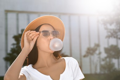 Photo of Beautiful woman in stylish sunglasses blowing gum outdoors on sunny day, space for text