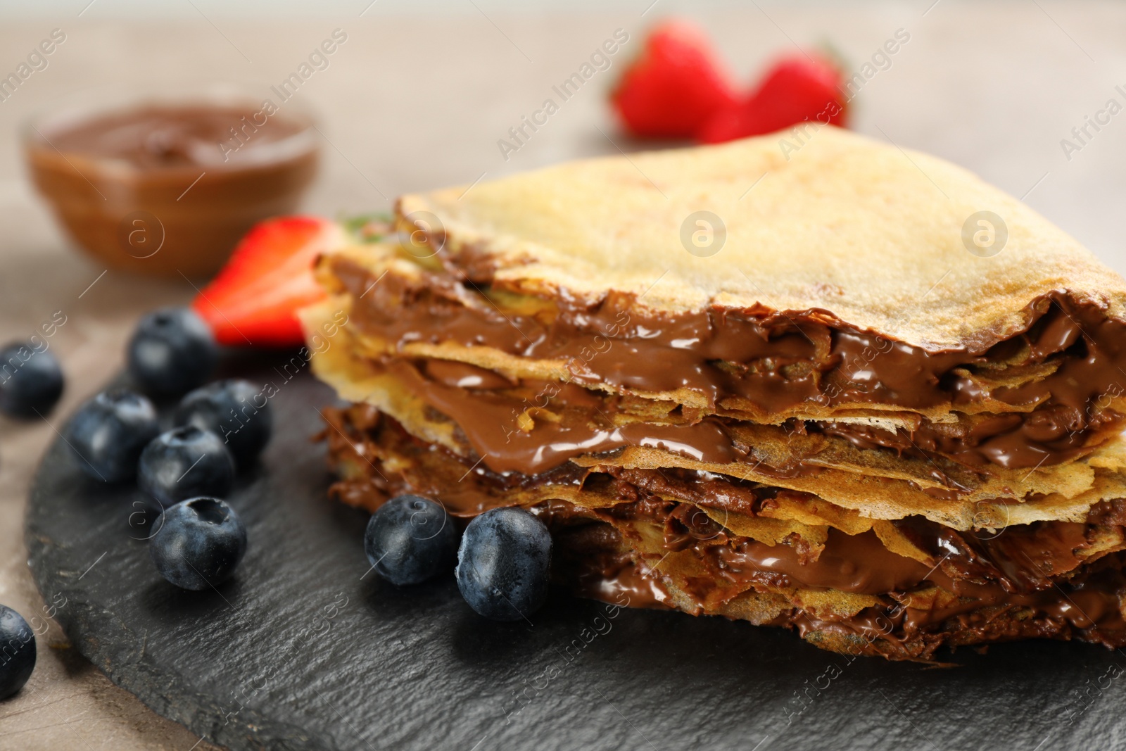 Photo of Delicious thin pancakes with chocolate paste and berries on slate plate, closeup