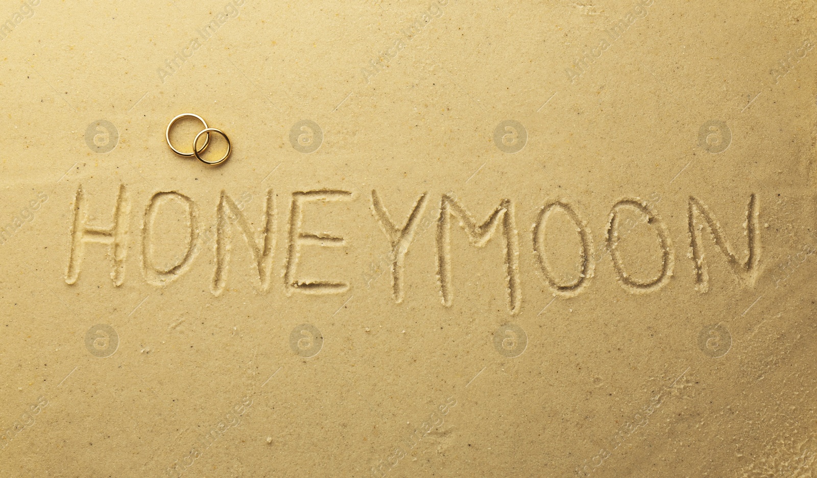 Photo of Word Honeymoon written on sand and two golden rings, top view