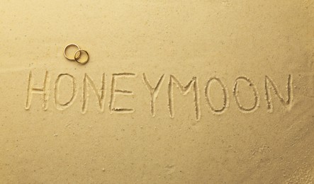 Photo of Word Honeymoon written on sand and two golden rings, top view