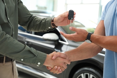 Photo of Young salesman shaking hands with customer and giving car key in modern auto dealership, closeup