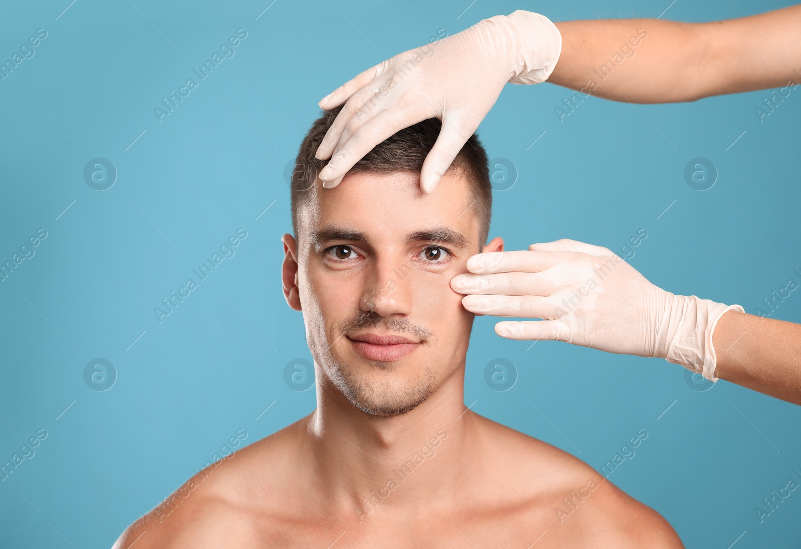 Photo of Doctor examining man's face for cosmetic surgery on light blue background