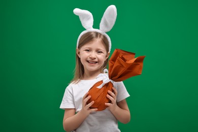 Photo of Easter celebration. Cute girl with bunny ears holding wrapped gift on green background