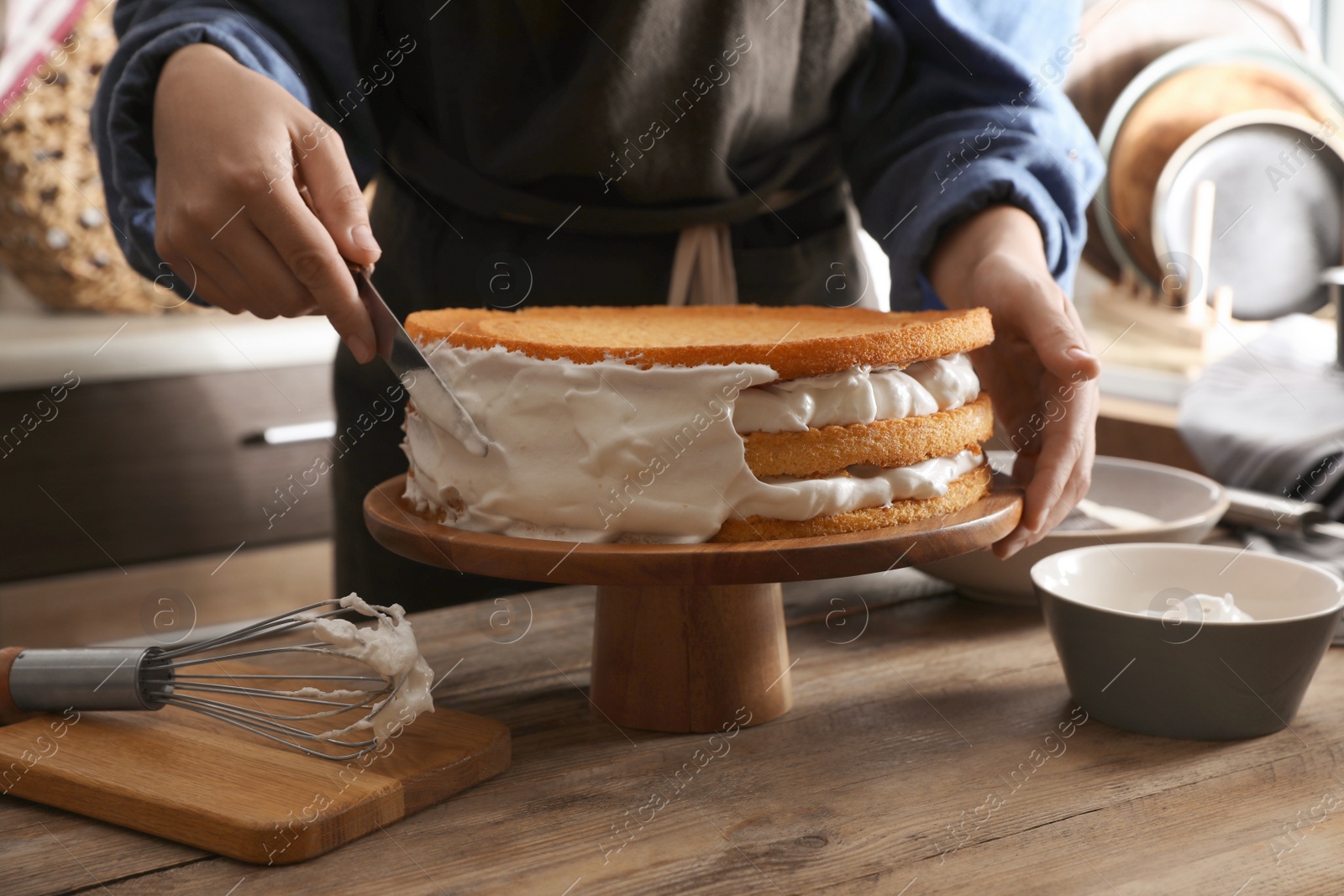 Photo of Woman smearing sides of sponge cake with cream at wooden table, closeup