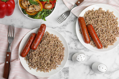 Photo of Tasty buckwheat porridge with sausages on white marble table, flat lay