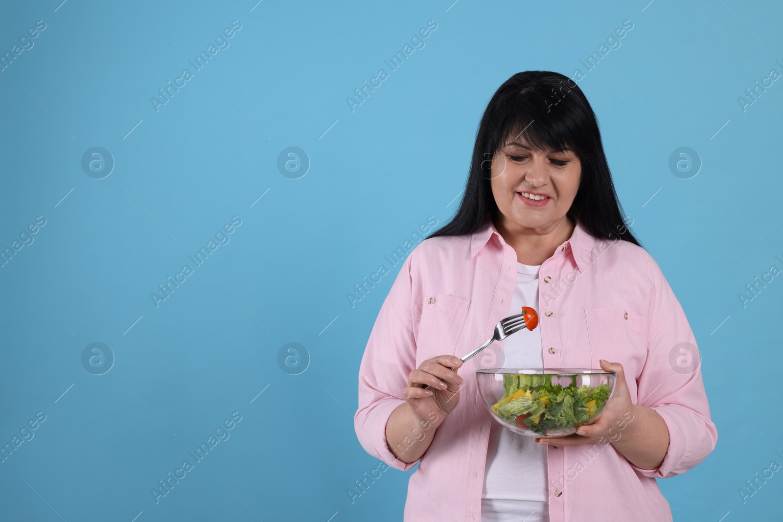 Photo of Beautiful overweight woman eating salad on light blue background, space for text. Healthy diet