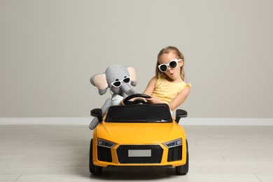 Photo of Cute little girl with toy elephant driving children's car near grey wall indoors