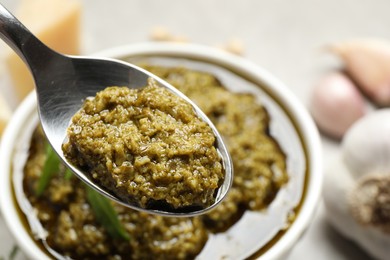 Photo of Spoon of tasty arugula pesto near bowl with sauce, closeup