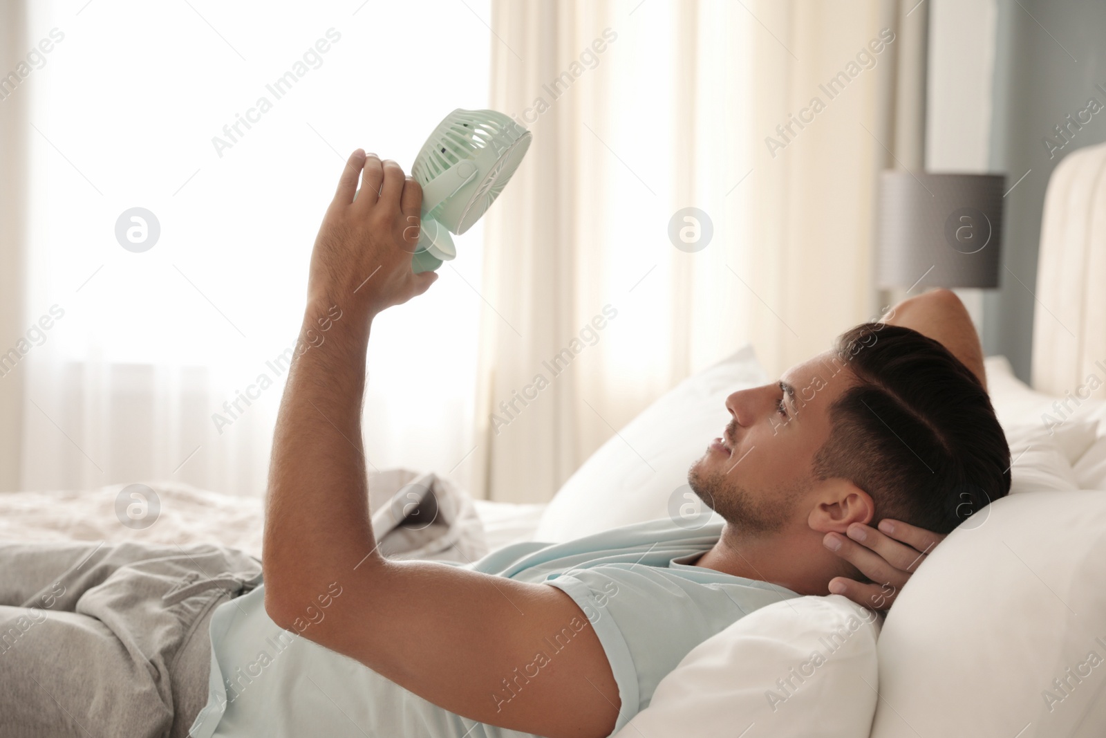 Photo of Man enjoying air flow from portable fan on bed in room. Summer heat