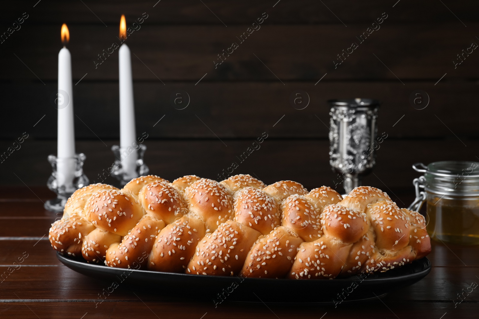 Photo of Homemade braided bread with sesame seeds, goblet and candles on wooden table. Traditional Shabbat challah