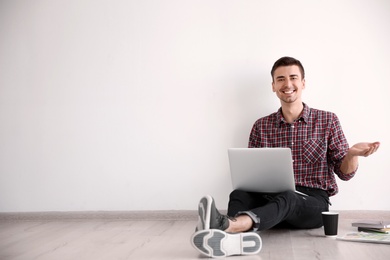 Young blogger with laptop sitting on floor against light wall