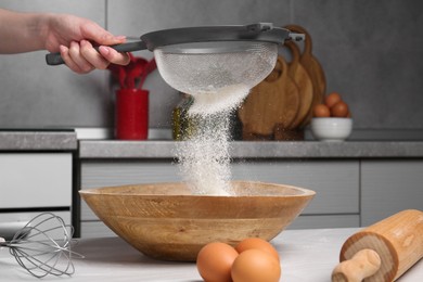 Photo of Woman sieving flour into bowl at white wooden table in kitchen, closeup