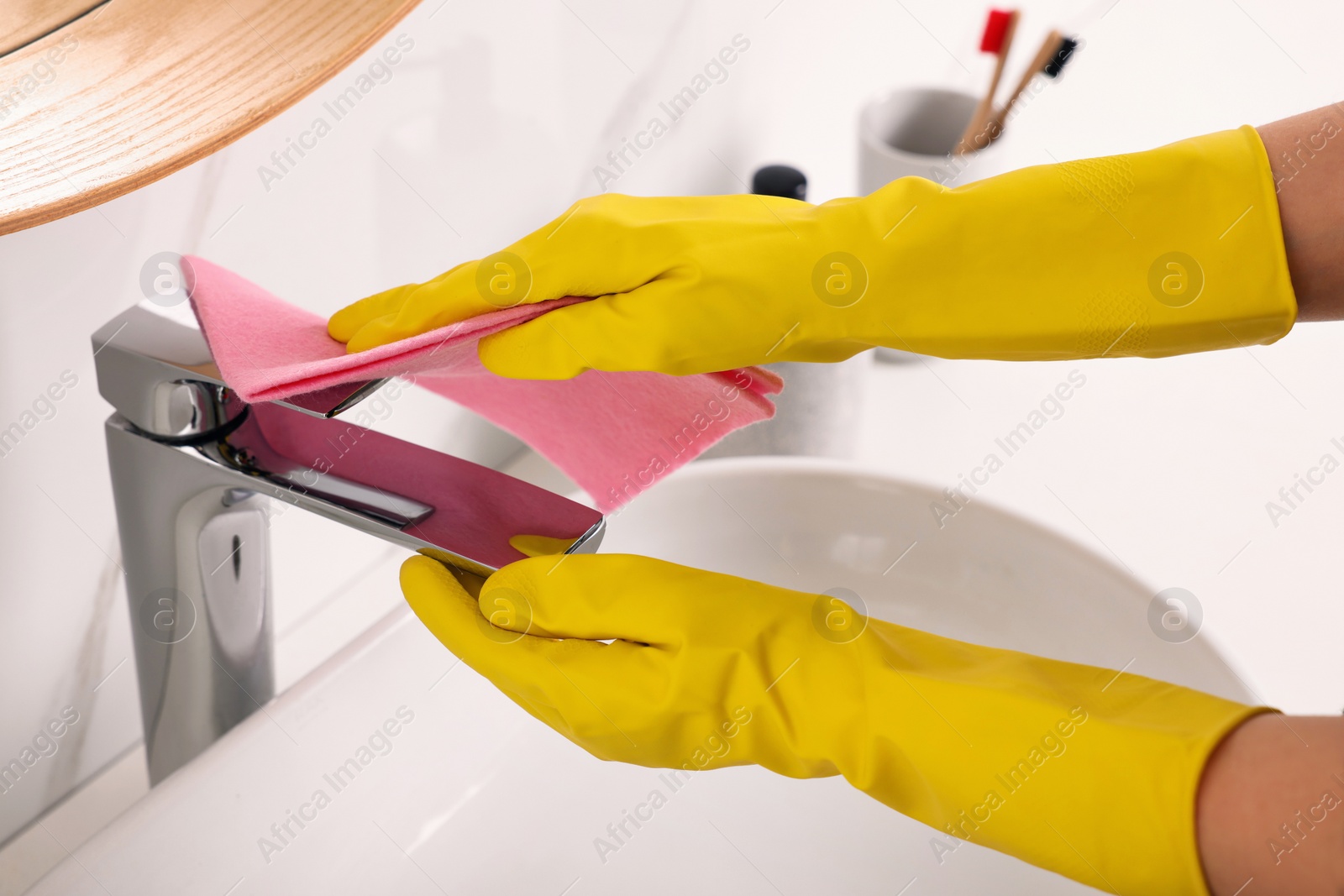 Photo of Woman in gloves cleaning faucet of bathroom sink with rag, closeup