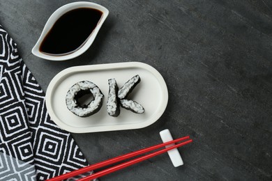 Photo of Word Ok made of sushi rolls, chopsticks and soy sauce on dark grey table, flat lay. Space for text