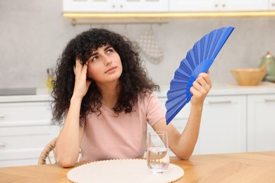 Young woman waving blue hand fan to cool herself at table in kitchen