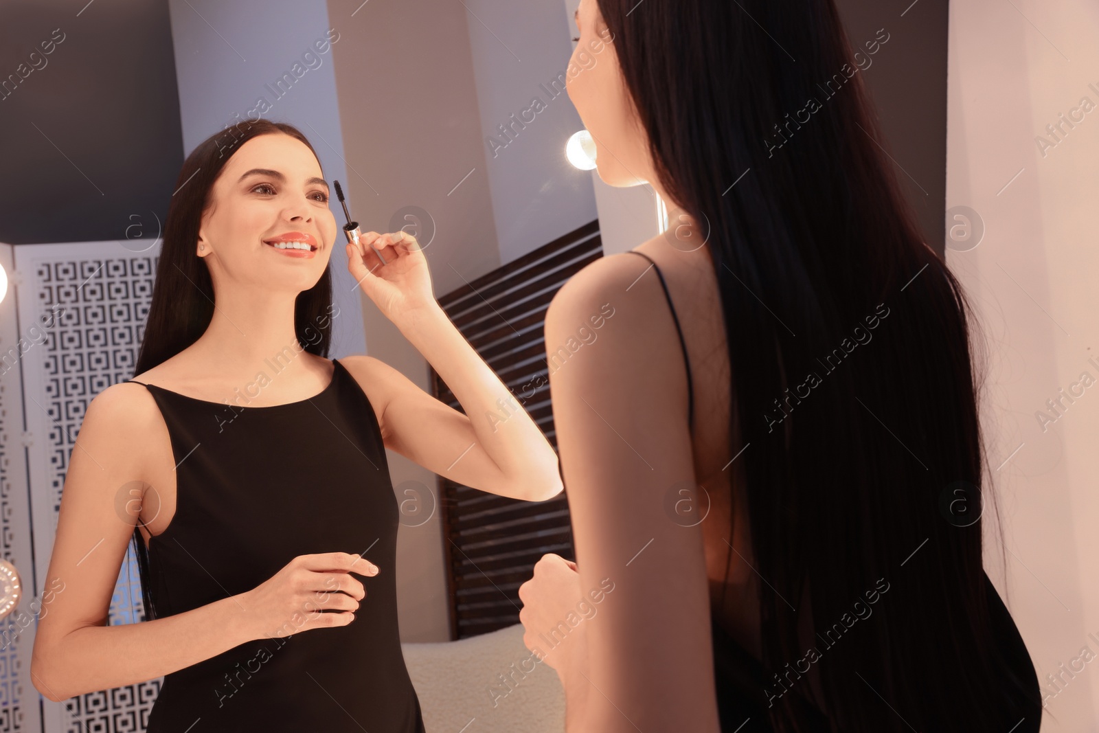 Photo of Beautiful young woman in elegant dress applying mascara near mirror indoors