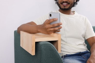 Photo of Man holding smartphone and listening to music on sofa with wooden armrest table at home, closeup