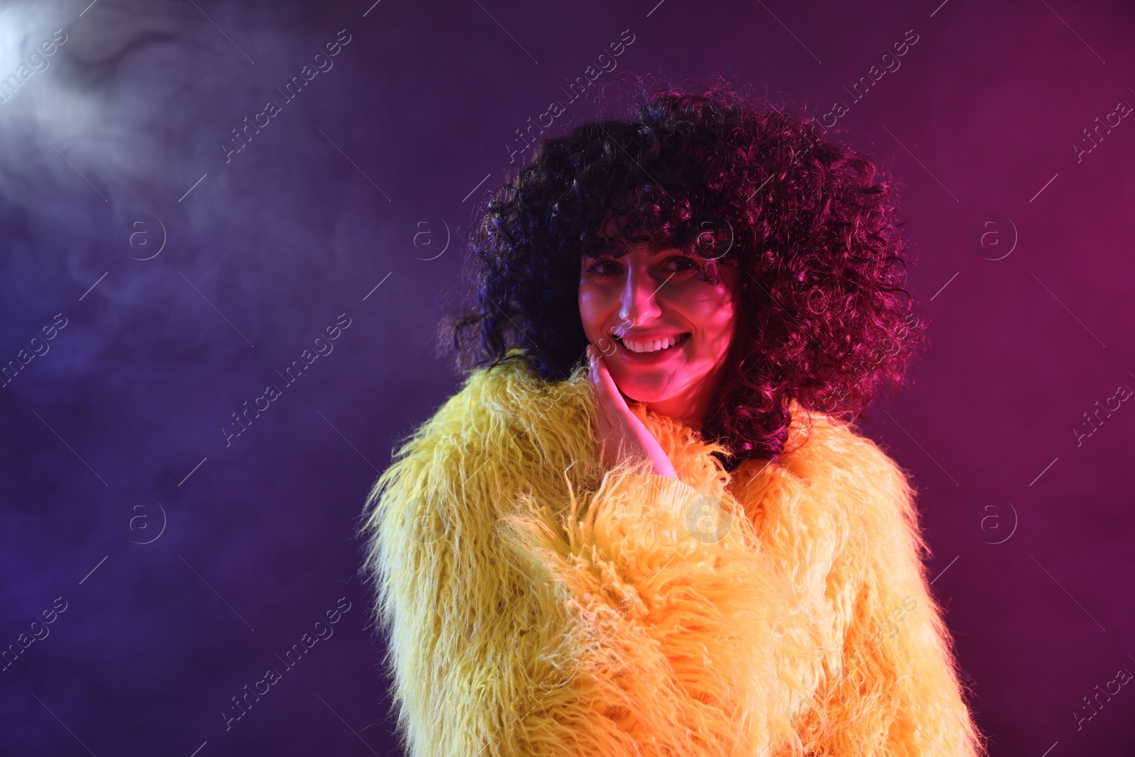 Photo of Beautiful young woman in yellow fur coat on color background in neon lights and smoke