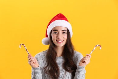 Beautiful woman in Santa Claus hat holding candy canes on yellow background