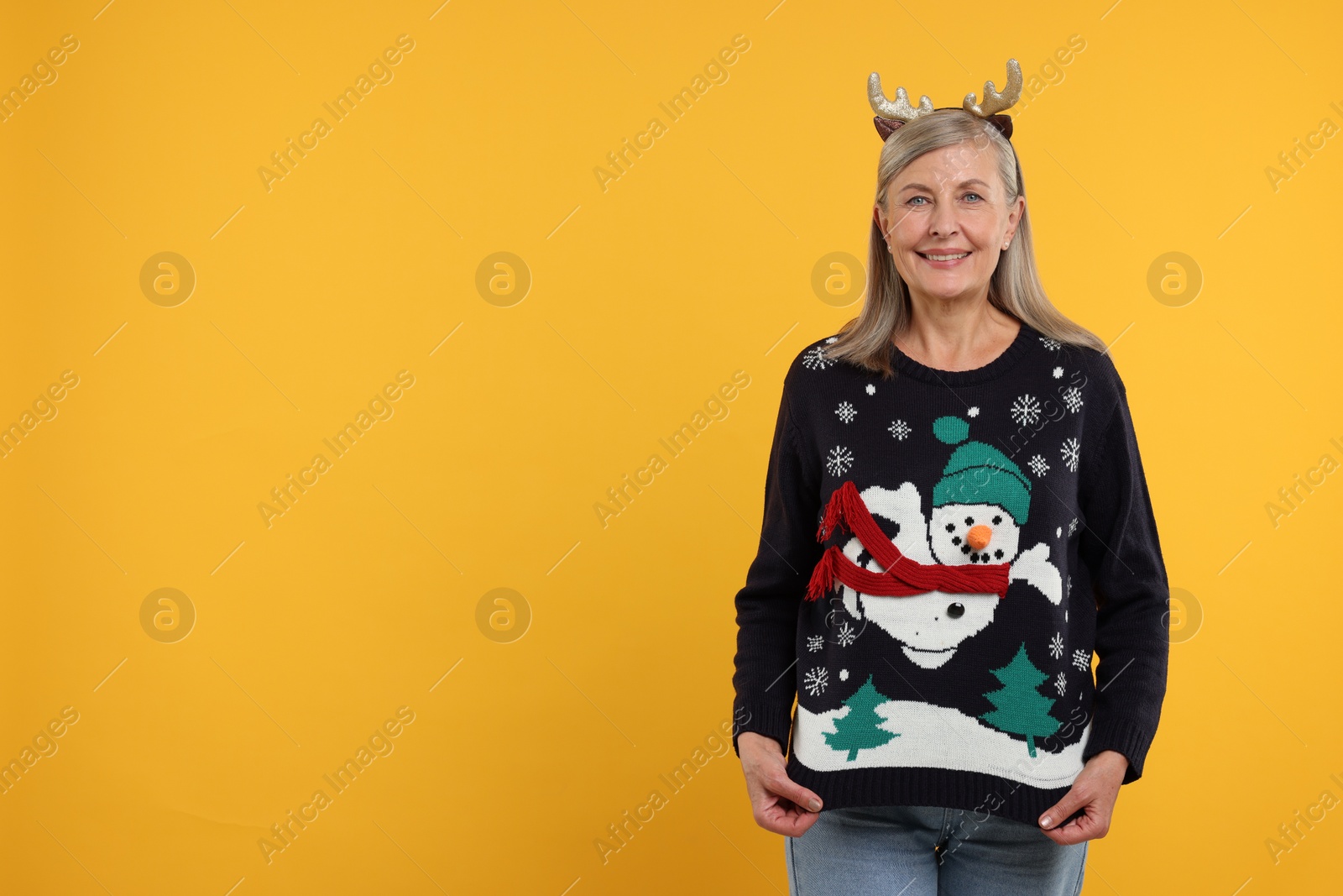 Photo of Happy senior woman in deer headband showing Christmas sweater on orange background. Space for text