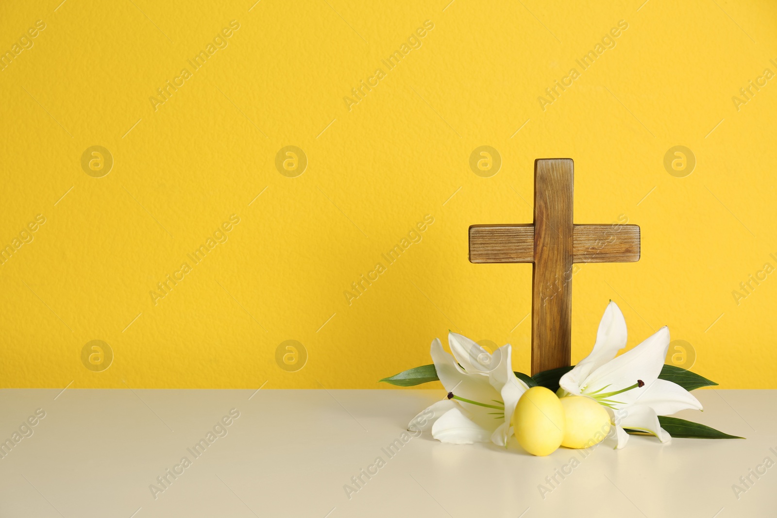 Photo of Wooden cross, Easter eggs and blossom lilies on table against color background, space for text