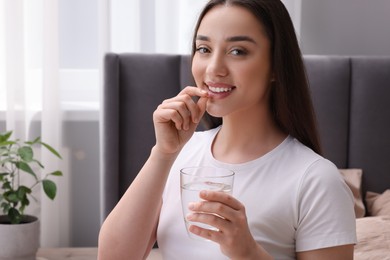 Photo of Beautiful young woman with glass of water taking pill at home, space for text