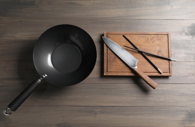 Black metal wok, knife, board and chopsticks on wooden table, top view